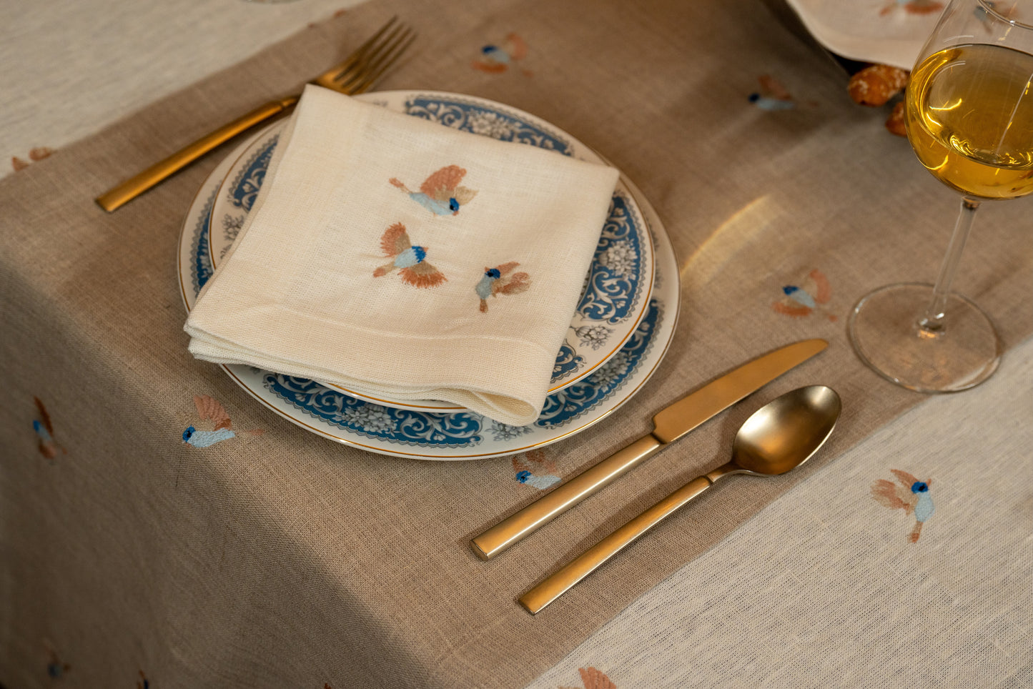 Dinner setting with plates, napkin, cutlery, and a glass on an Egyptian linen tablecloth with colorful bird embroidery.