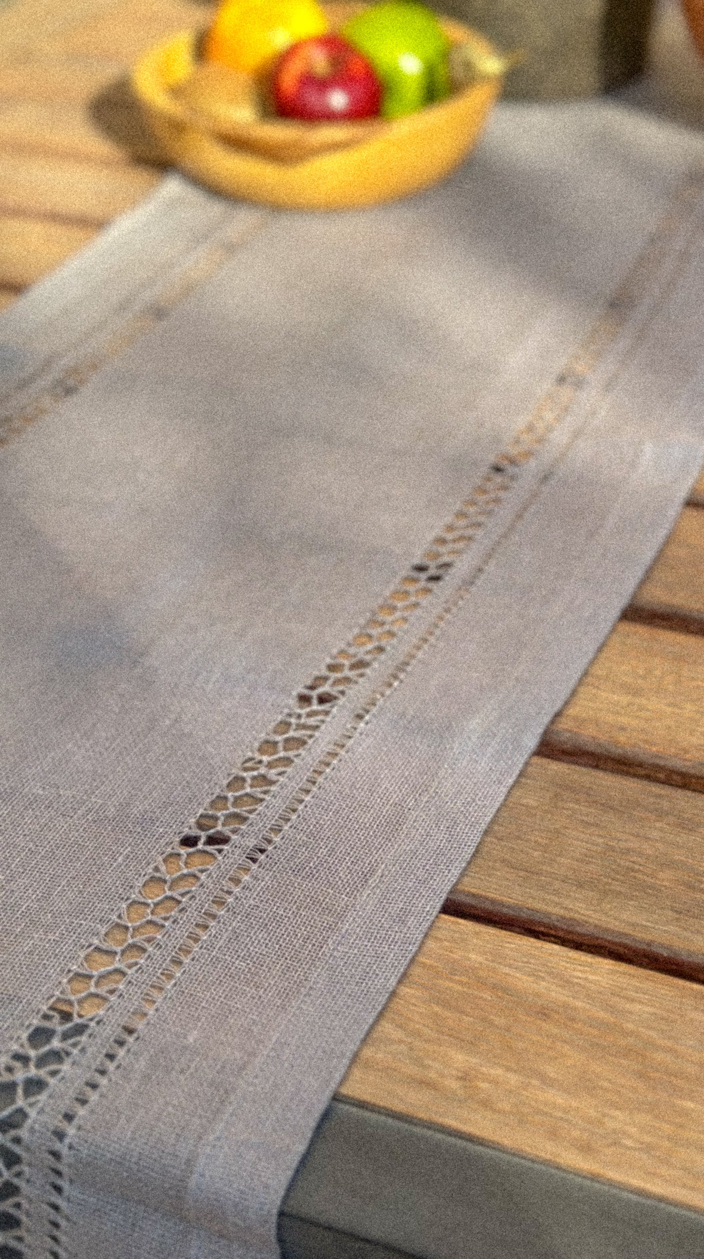 Gray handmade table runner with lace trim on a wooden table, with fruits in the background.