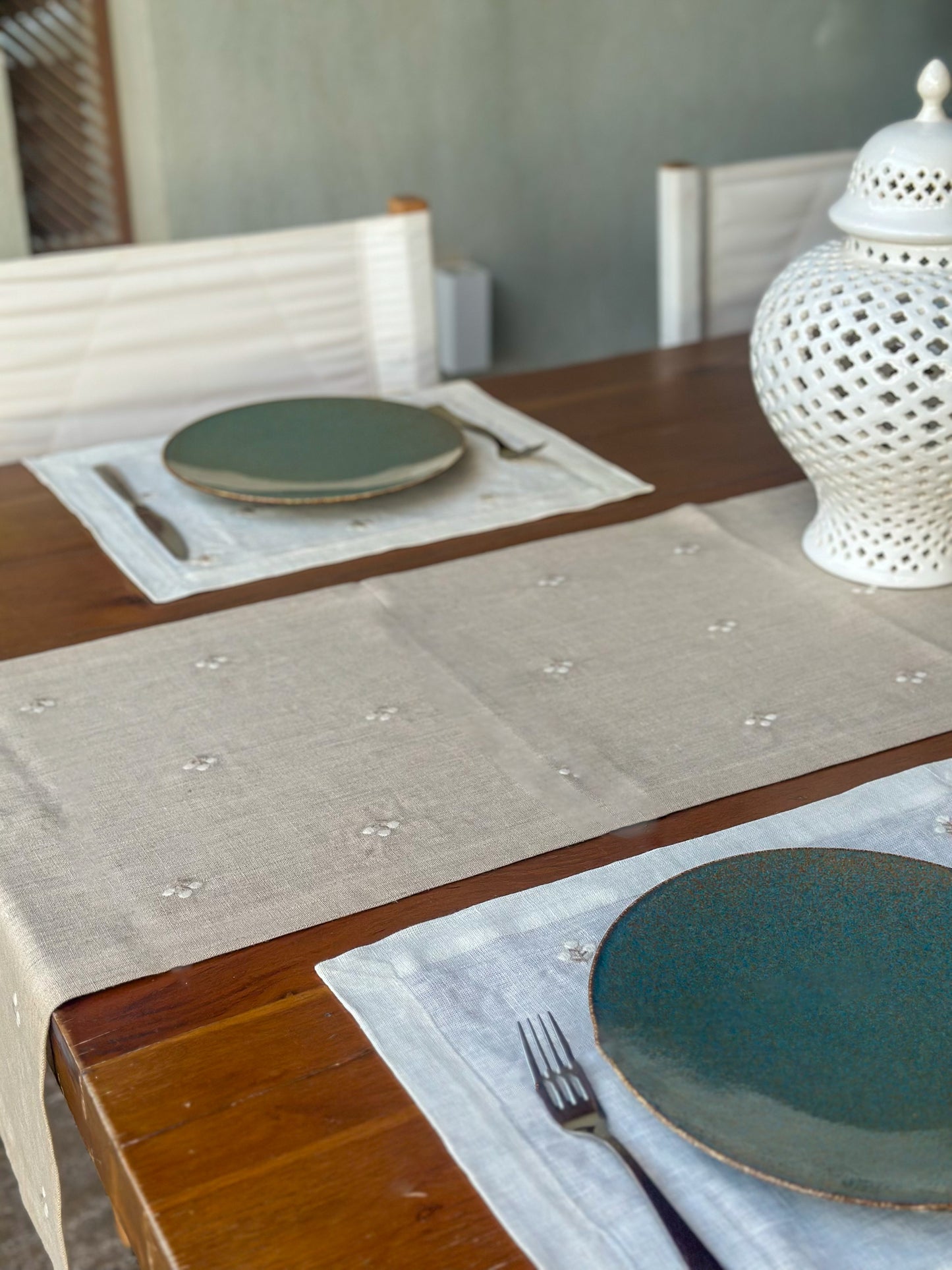 Dining table setting with embroidered linen runner, placemats, plates, cutlery, and a decorative lamp.