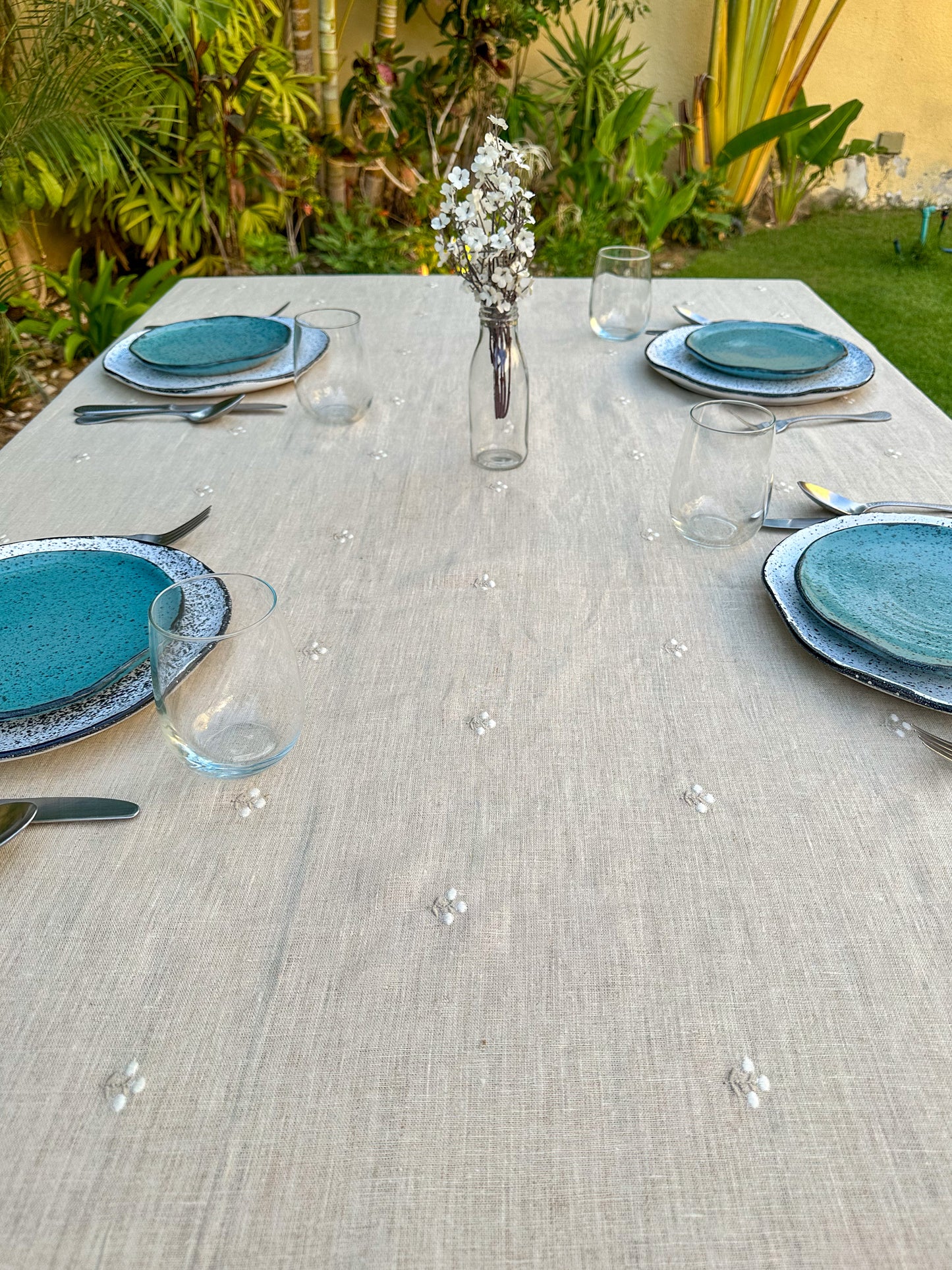 Dining table set with blue plates, silverware, and glasses on a beige embroidered Egyptian linen tablecloth outdoors.
