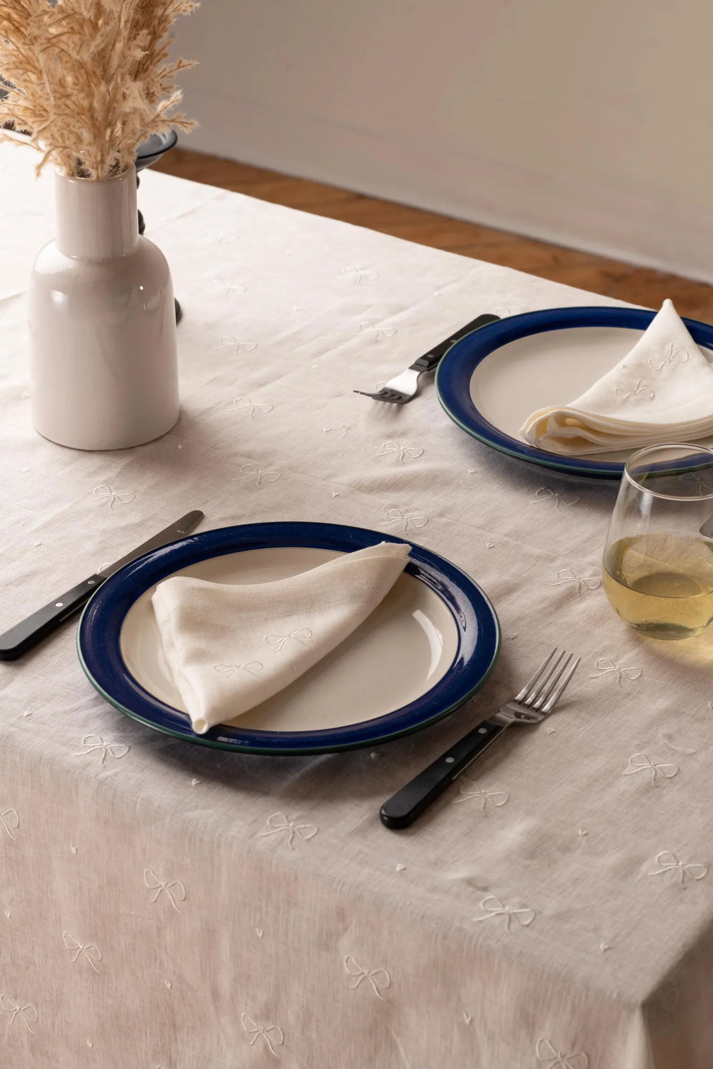 A white embroidered linen tablecloth on a table with two plates, blue trim, and a glass of wine in the foreground.