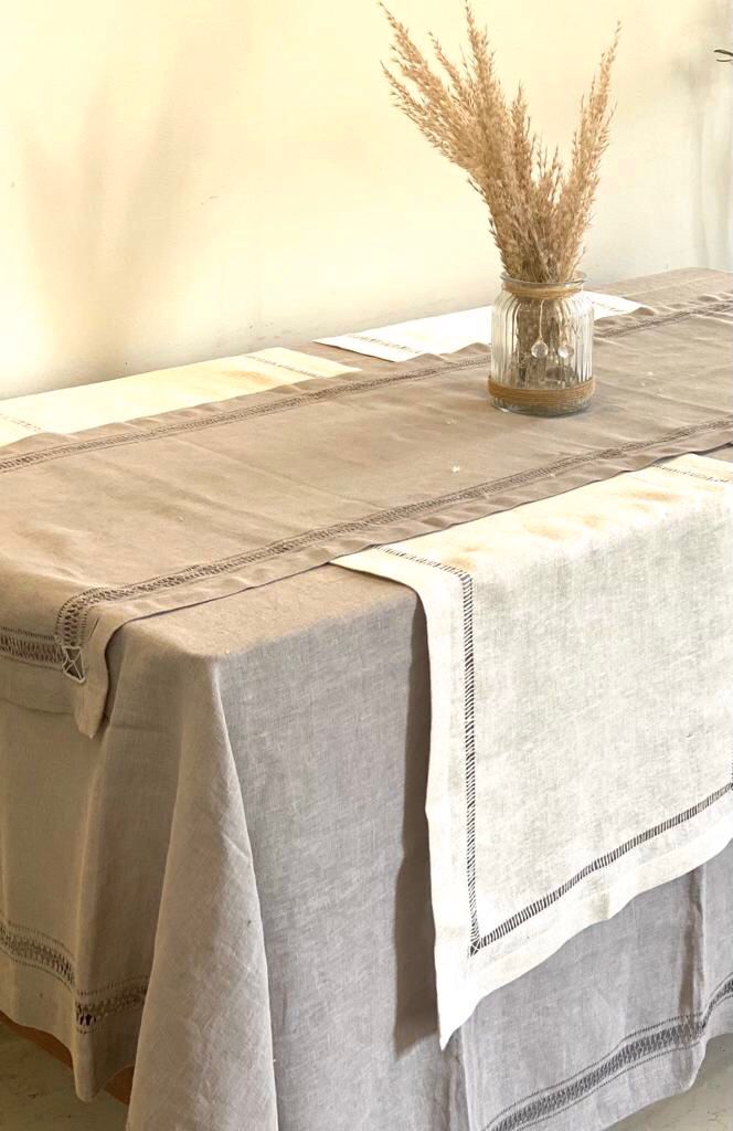 A beige handmade linen table runner with a decorative edge is displayed on a table, with a vase of dried plants and a white bowl in the background.