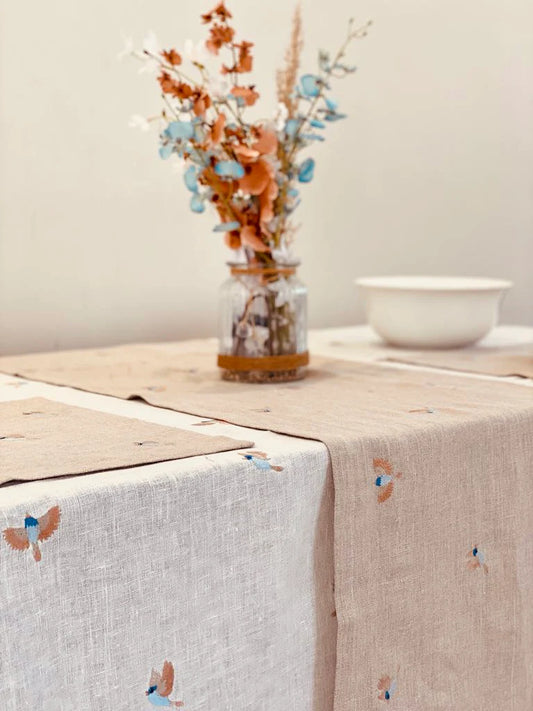 Table setting with embroidered tablecloth, linen runner, vase with dried flowers, and bowl on a neutral background