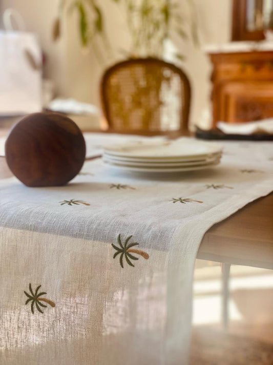 A beige Egyptian linen table runner with green palm tree embroidery laid out on a dining table with plates and a wooden bowl in the background.