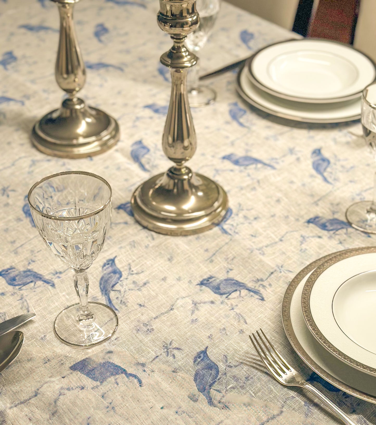 Dining table setting with silver candlesticks, glasses, and plates on an Egyptian linen tablecloth with blue bird embroidery.