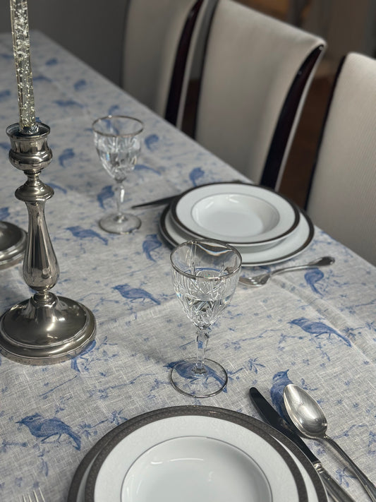 Dining table setting with plates, glasses, and silverware on a patterned Egyptian linen with blue bird print.