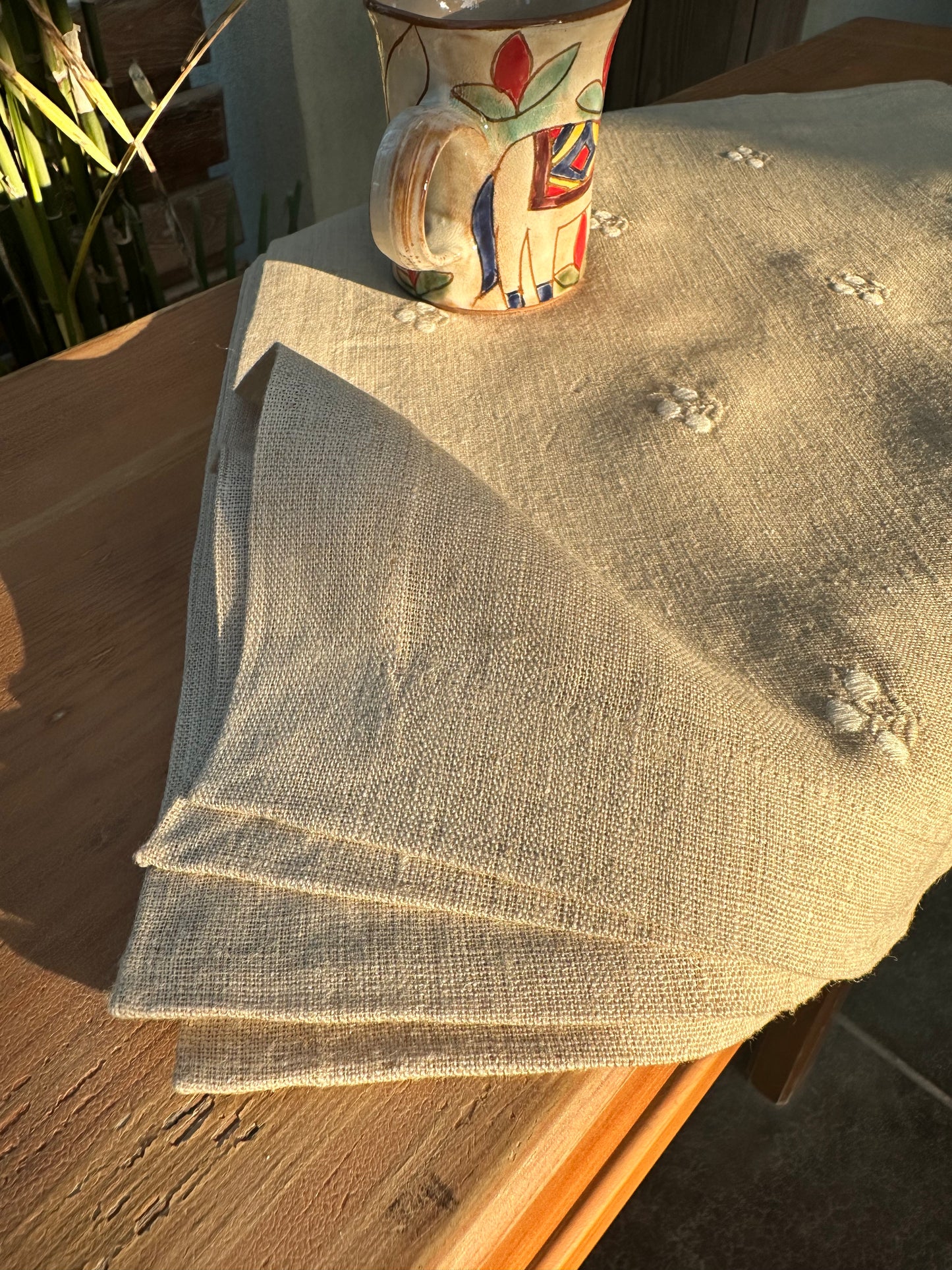 Stack of beige Egyptian linen placemats on a wooden surface with a colorful ceramic cup in the background.