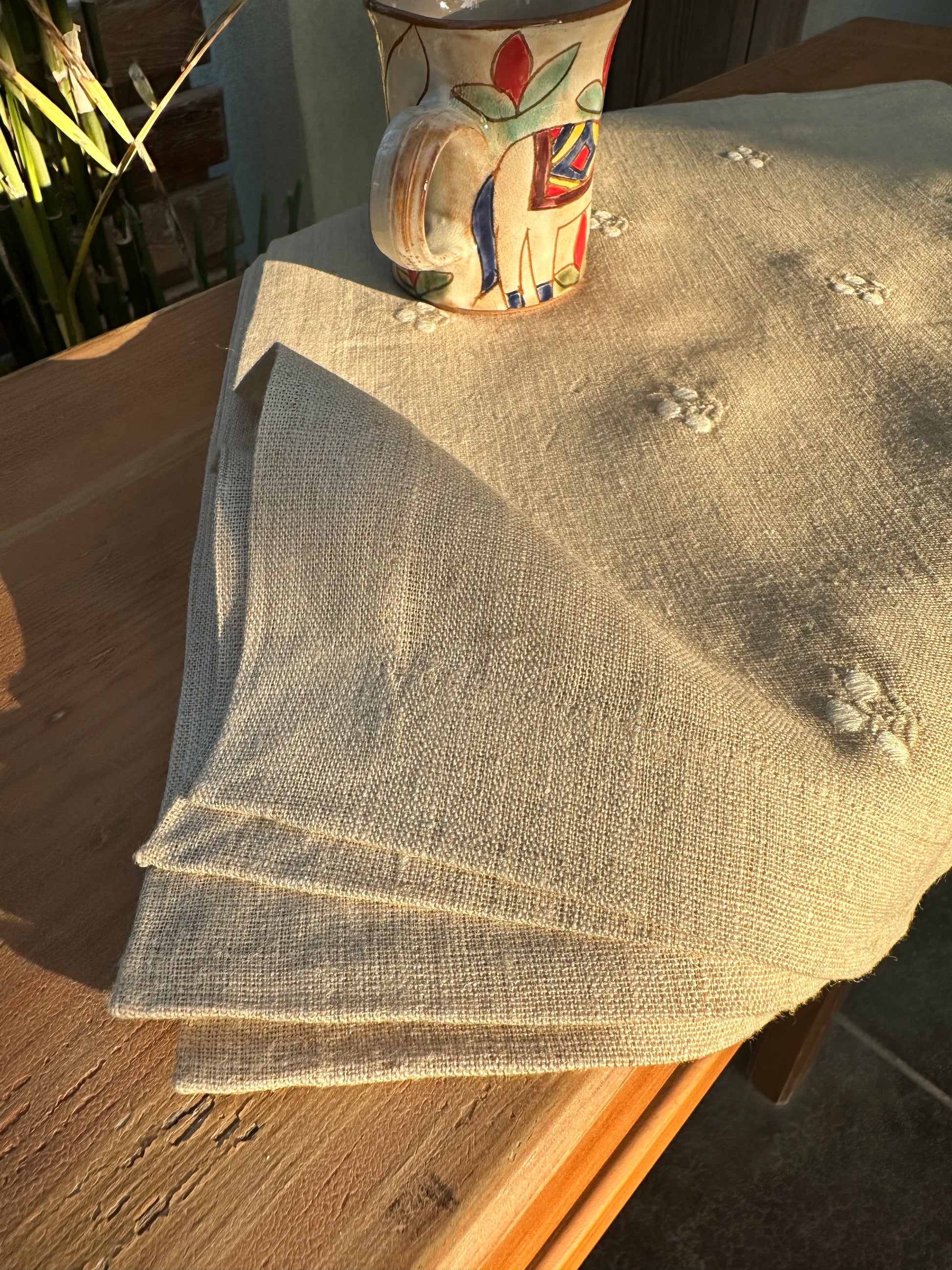 Stack of beige Egyptian linen placemats on a wooden surface with a colorful ceramic cup in the background.