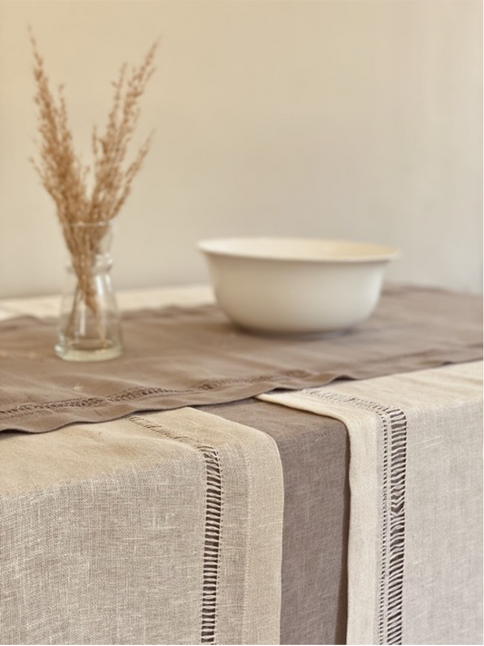 A beige handmade linen table runner with a decorative edge is displayed on a table, with a vase of dried plants and a white bowl in the background.